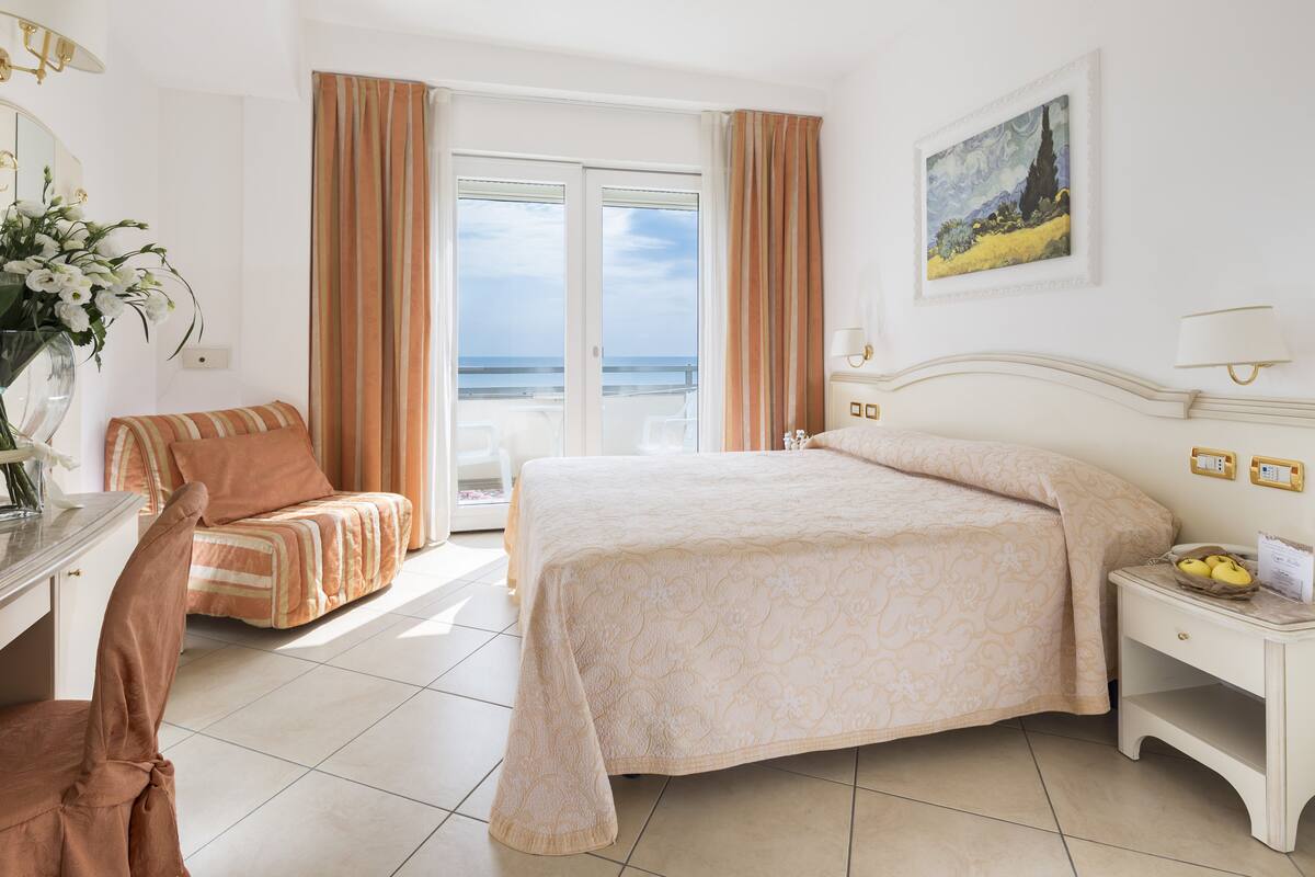 A hotel room with a bed, a chair, a nightstand, and a view of the ocean through the window.