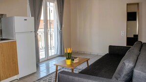 Ein Wohnzimmer mit einer Couch, einem kleinen Holztisch mit Büchern und Stiften, einem Kühlschrank und Blick durch ein Fenster mit Vorhängen.