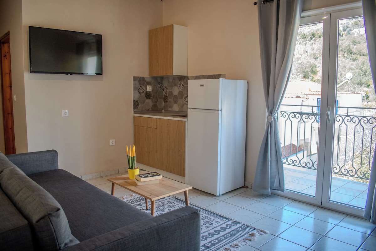 A living room with a sofa, a coffee table with books and pencils, a TV mounted on the wall, a kitchen with a refrigerator and cabinets, and a balcony with a railing.
