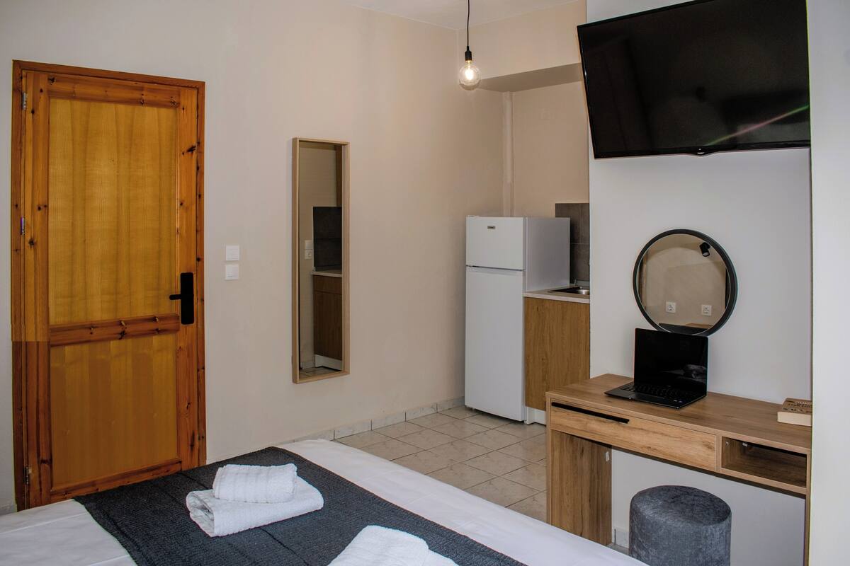 A room with a bed, a desk with a laptop, a TV, a mirror, a refrigerator, and a wooden door.