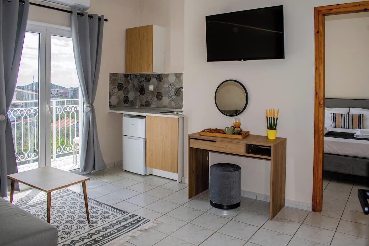 A compact room with a kitchenette, a small desk, a trash bin, a bed, and a balcony with curtains.