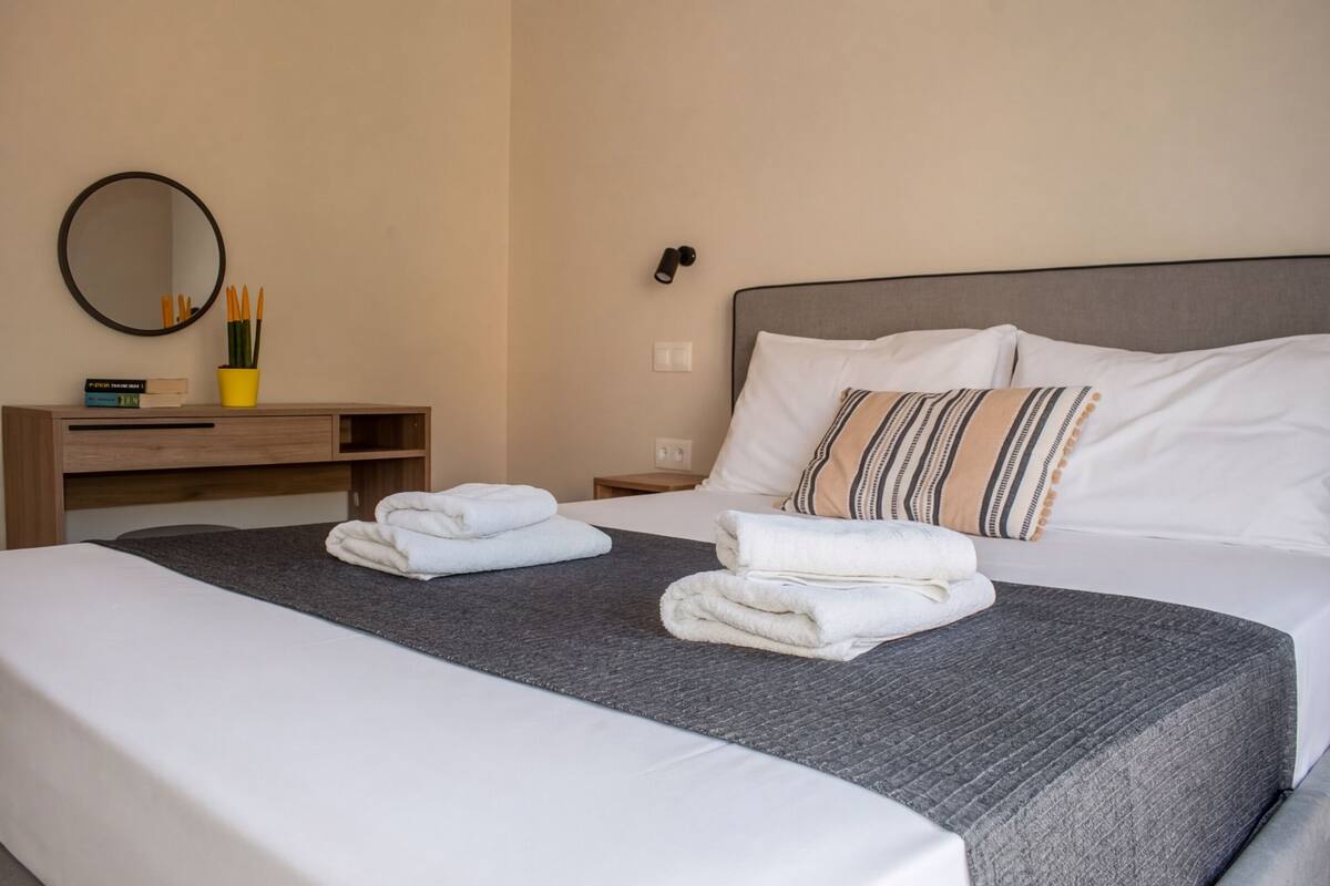 A hotel room with a bed, a bedside table with a mirror, a lamp, and a small shelf with books and a plant.