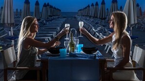 Two people toasting at a table with a bottle and glasses on a beach with rows of umbrellas. - Hotel Mocambo (Cervia)