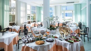 A dining area with round tables covered in white tablecloths, featuring a variety of food items and a central white column. - Hotel Mocambo (Cervia)