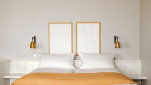 Una cama con cabecera blanca, dos lámparas fijadas a la pared y una mesita de noche blanca.