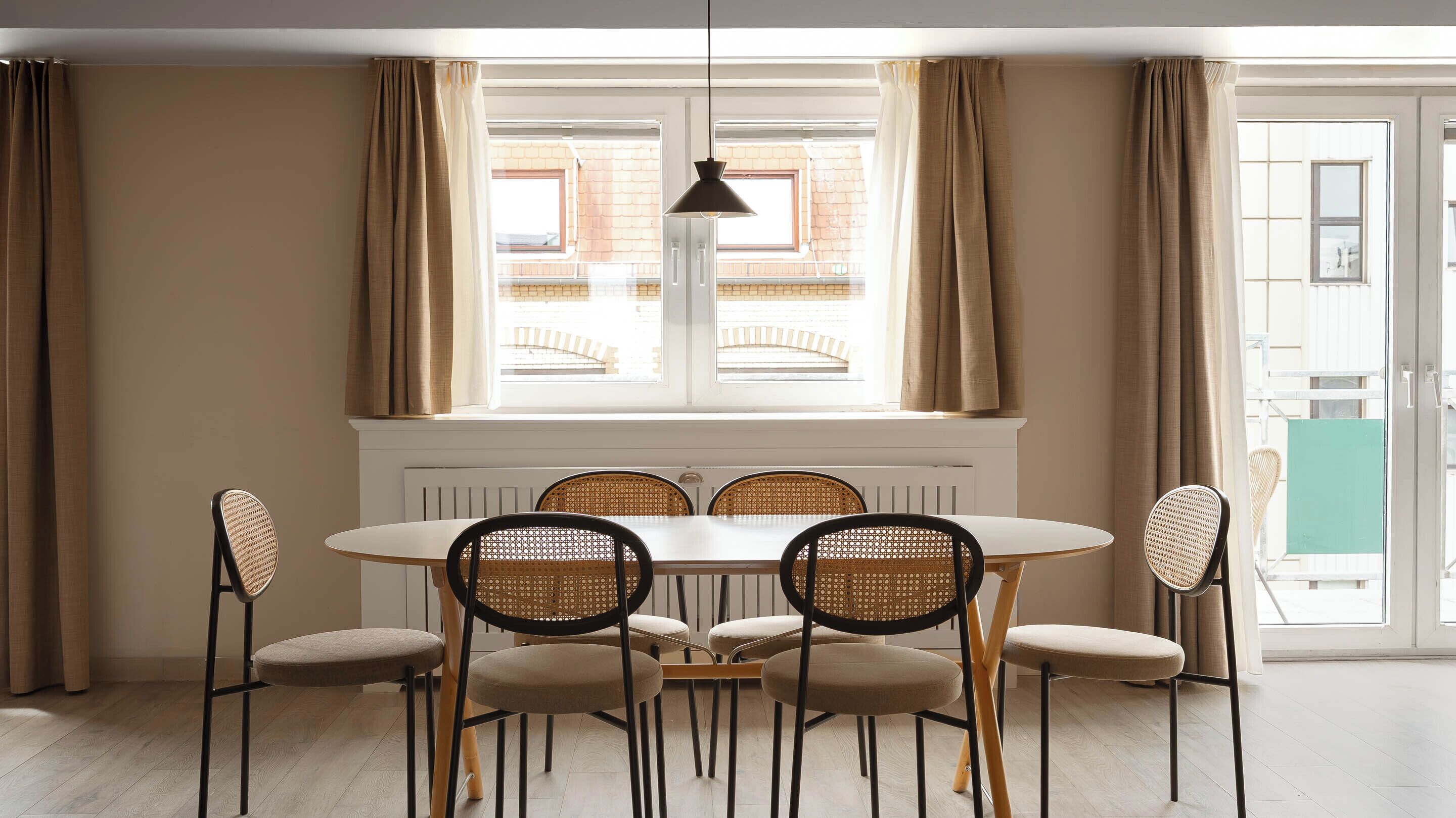 A dining area with a round table and six chairs, a hanging pendant light, and a view through a window with curtains.