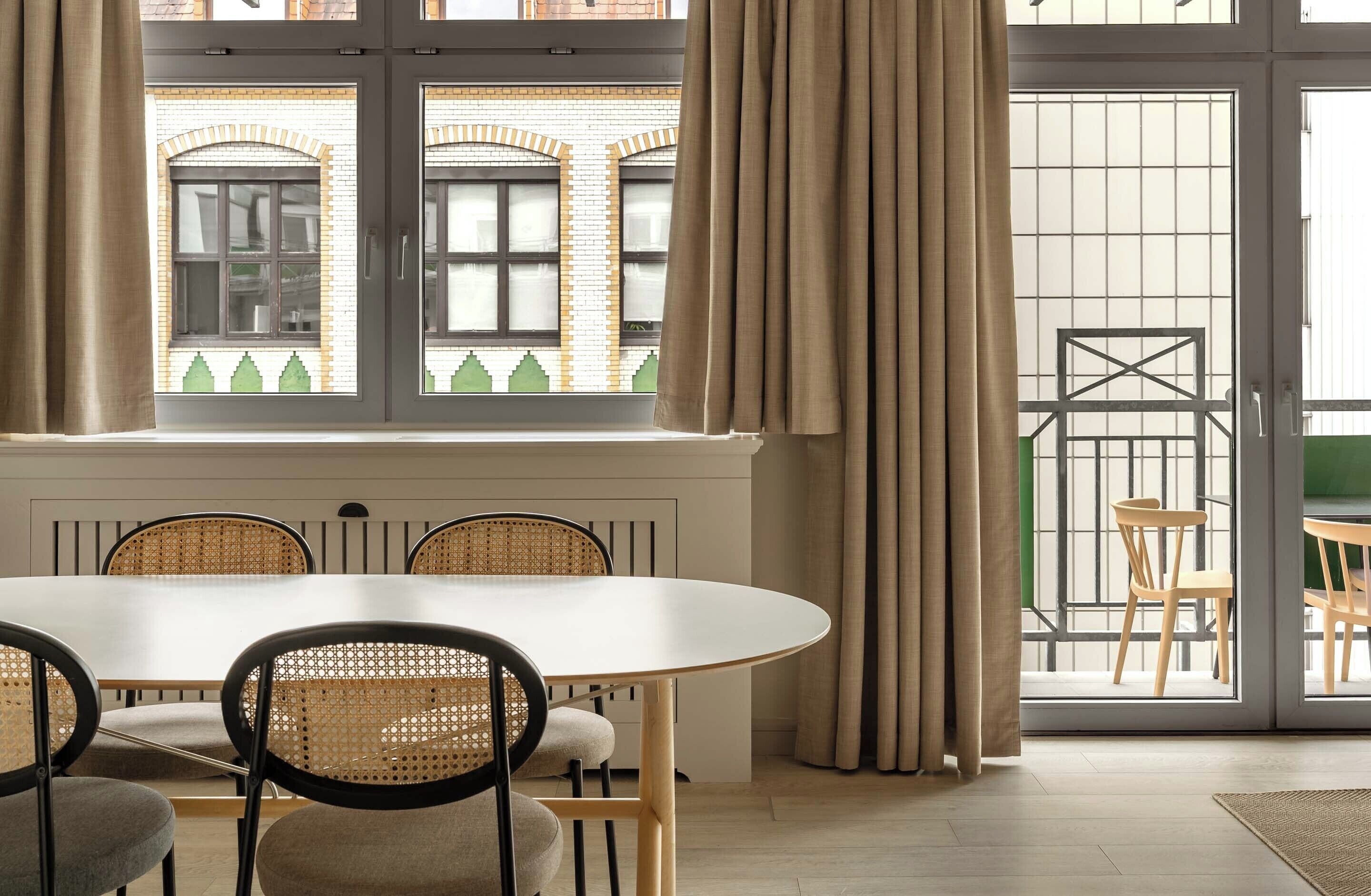 Un comedor con una mesa redonda y sillas, una ventana con cortinas, y vistas a un edificio con ladrillo y un balcón.