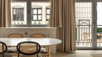 Un comedor con una mesa redonda y sillas, una ventana con cortinas, y vistas a un edificio con ladrillo y un balcón.