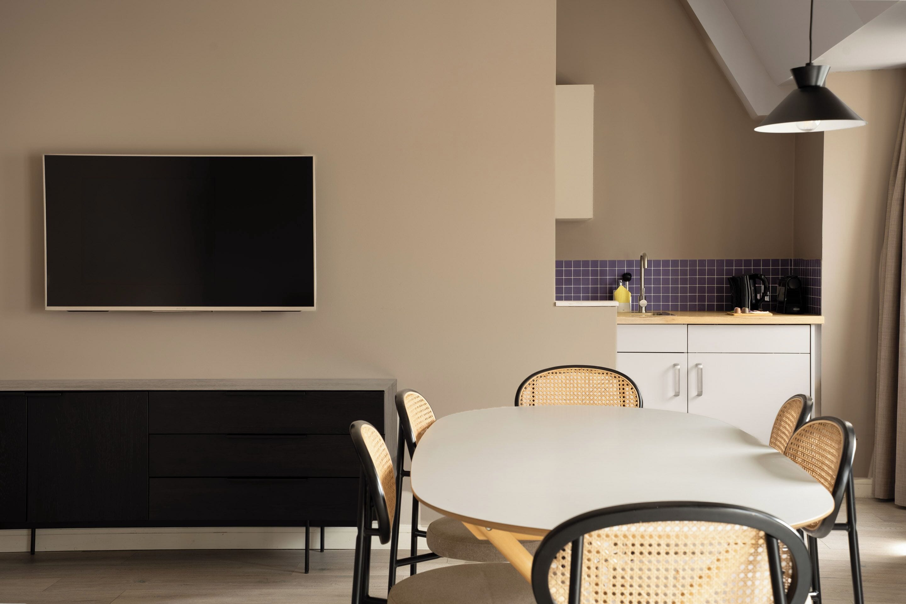 Una cocina moderna con comedor, un televisor de pantalla plana montado en la pared, un armario negro y sillas de mimbre alrededor de una mesa blanca.