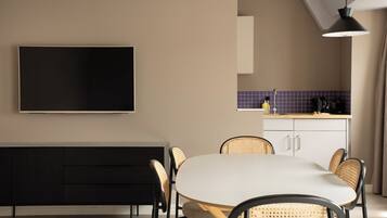 Una cocina moderna con comedor, un televisor de pantalla plana montado en la pared, un armario negro y sillas de mimbre alrededor de una mesa blanca.