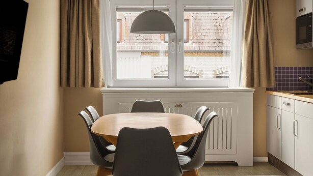 Un comedor con una mesa redonda, sillas, una ventana con cortinas, un radiador y una zona de cocina con armarios y un microondas.