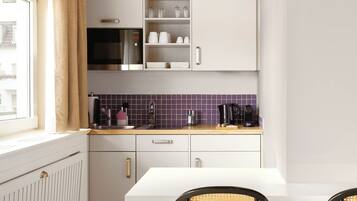 Cocina compacta con alacenas blancas, microondas integrado, cafetera y ventana con cortinas.