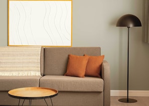 Una sala de estar con un sofá, dos almohadas, una mesita redonda, una lámpara de pie y una obra de arte abstracta enmarcada en la pared.