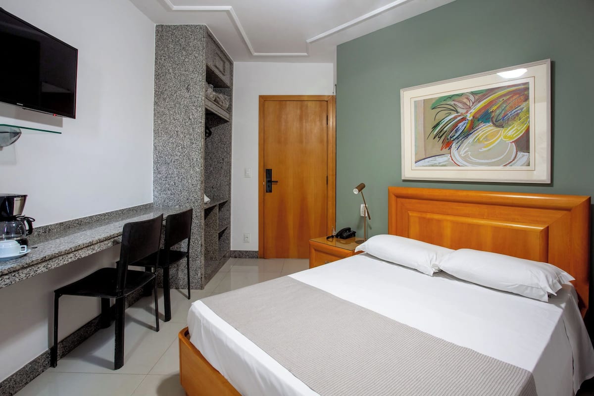 A hotel room with a bed, a desk with a chair, a coffee maker, a TV, and a painting on the wall.