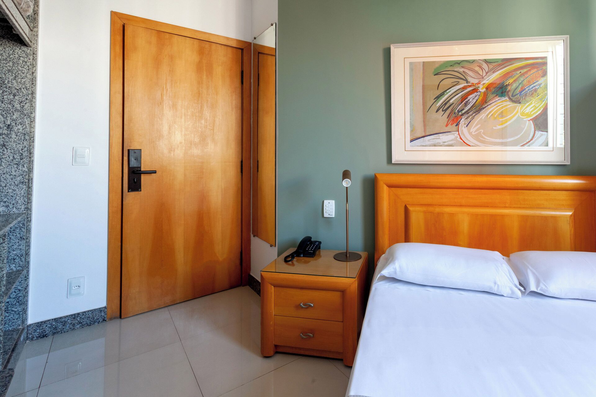 A hotel room with a wooden door, a bedside table with a lamp, a phone, and a bed with white linens.