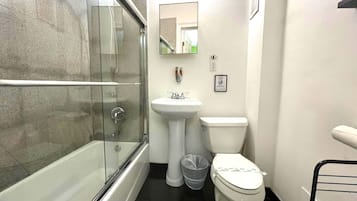 A bathroom with a glass-enclosed shower, a pedestal sink, a toilet, and a trash bin.