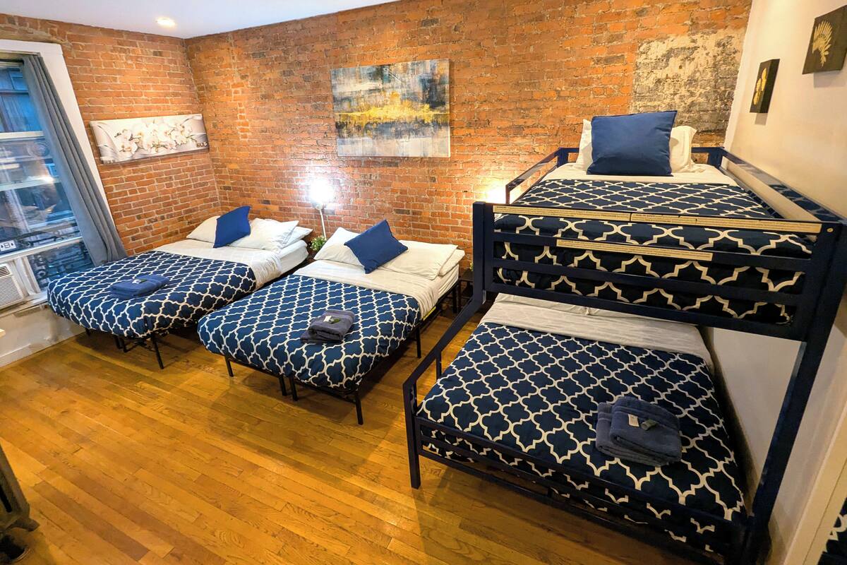 Ein Zimmer mit Ziegelwand, zwei Betten mit gemusterten Bettbezügen, einem Fenster mit Vorhängen und einem Bild an der Wand.