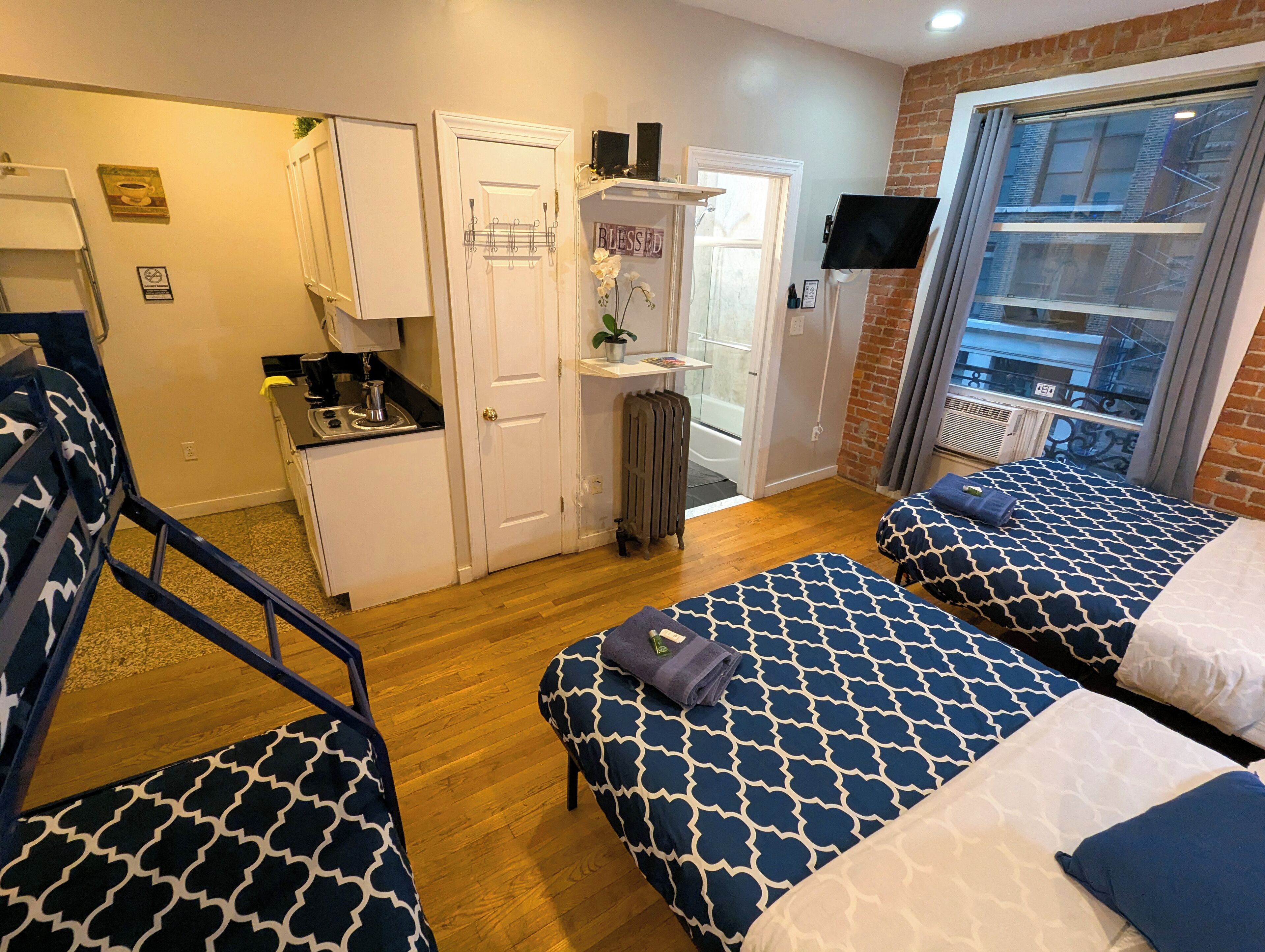 Une chambre d’hôtel avec deux lits, une kitchenette, un radiateur et une vue sur la ville grâce à une grande fenêtre.