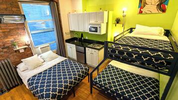 A room with two bunk beds, a kitchenette, a brick wall, and a window with blinds.