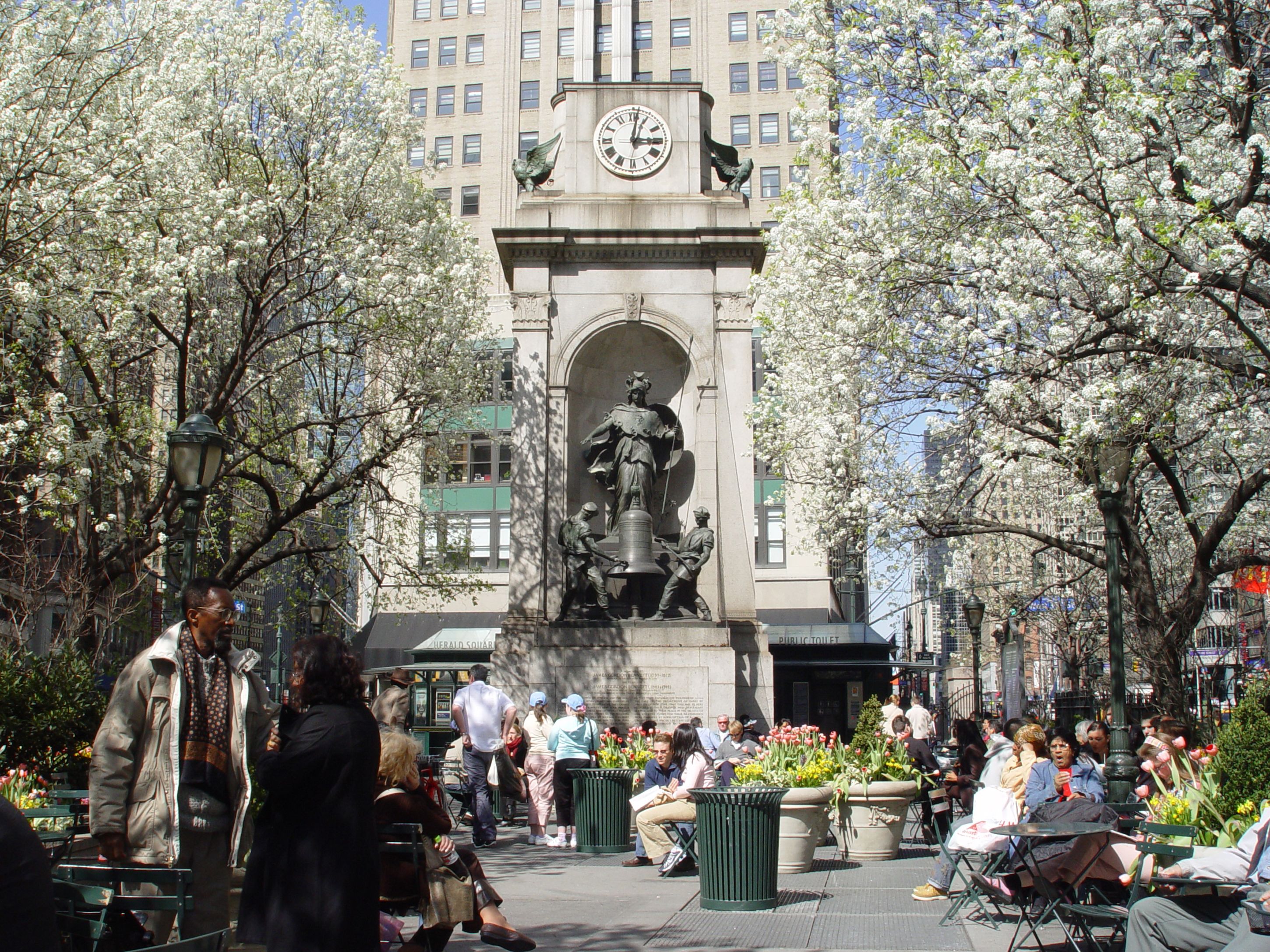 A public square with a clock tower, statues, and people sitting on benches.