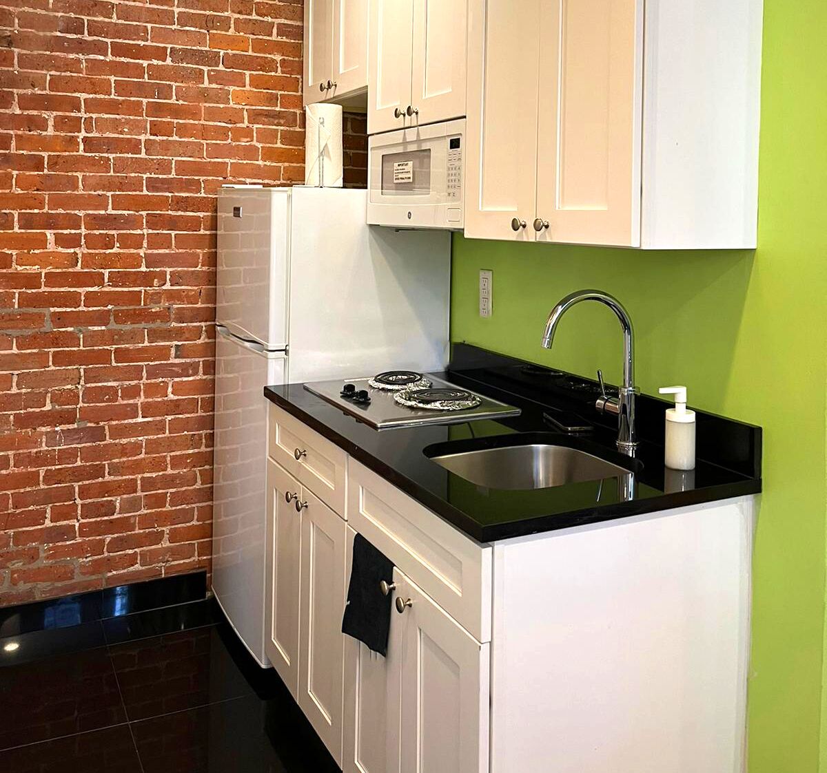 A compact kitchen with white cabinets, a black countertop, a stainless steel sink, and a brick wall.