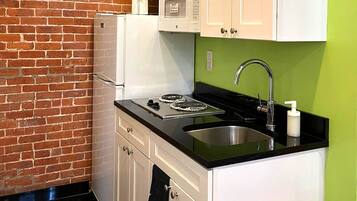 A compact kitchen with white cabinets, a black countertop, a stainless steel sink, and a brick wall.