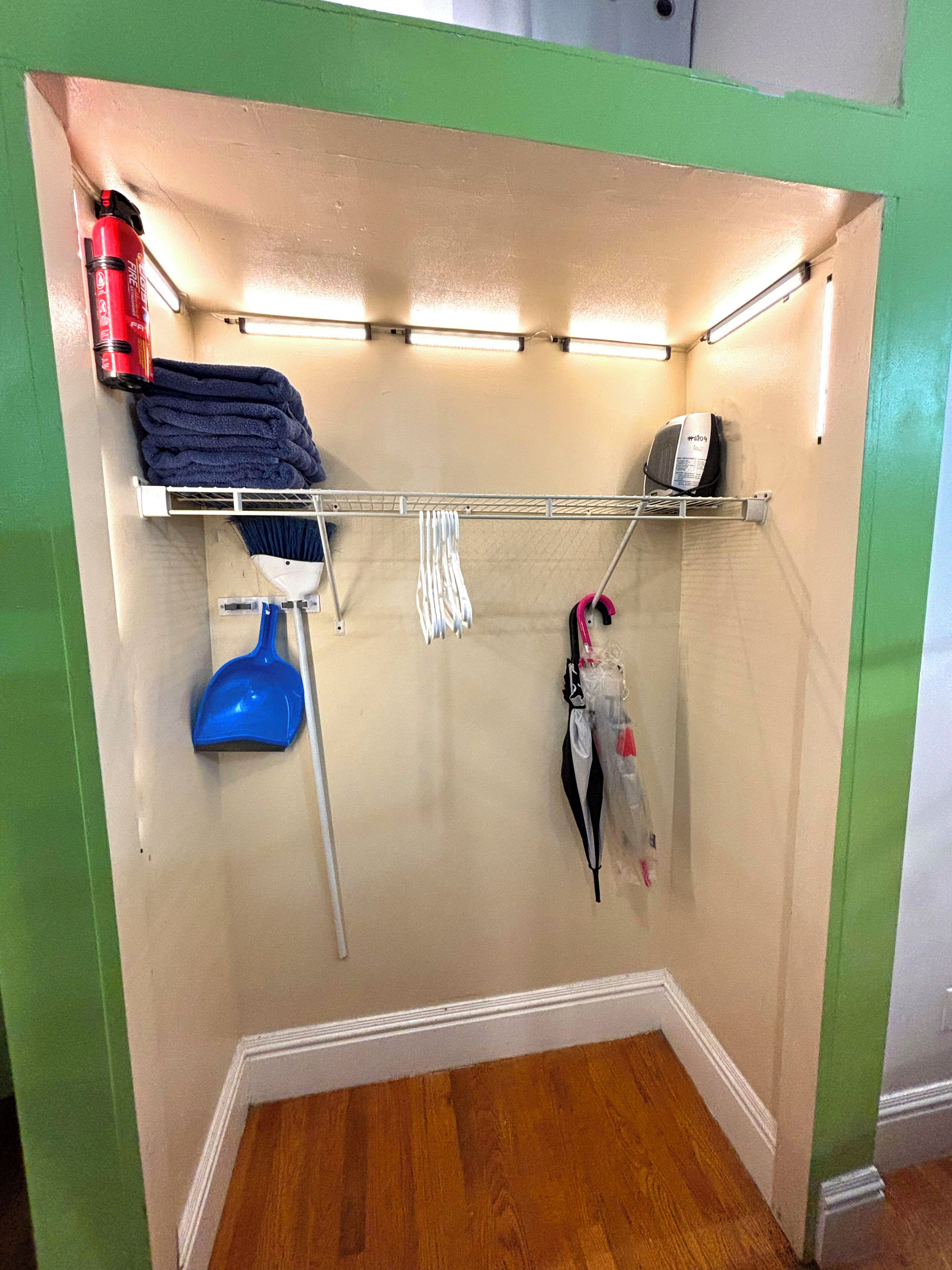 A corner closet with a metal rack holding towels, a blue dustpan, a white broom, and a red fire extinguisher.