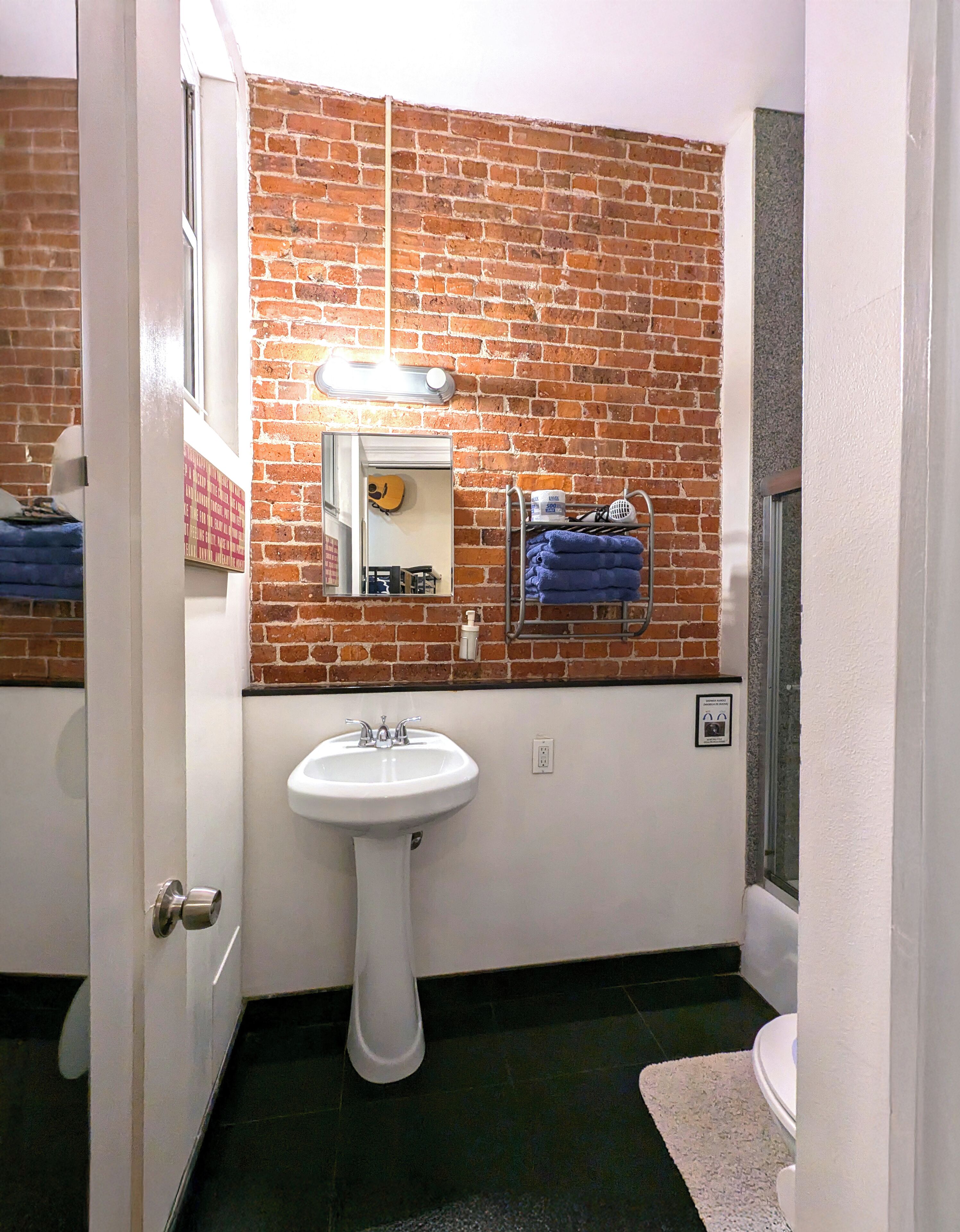 A bathroom with a white pedestal sink, a mirror, a towel rack with towels, a window, and a brick wall.