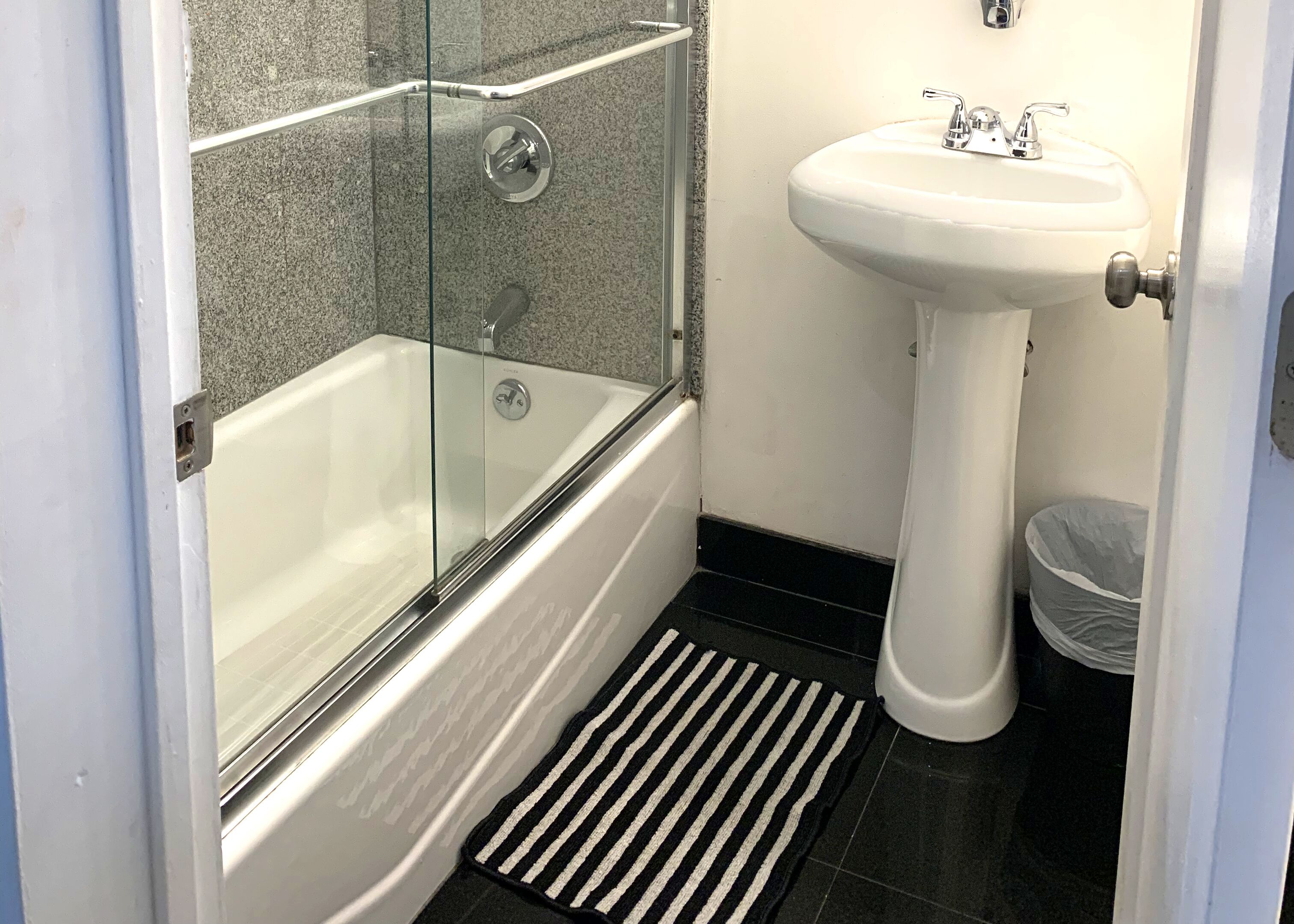 Un baño con ducha de vidrio, un lavamanos pedestal blanco y un felpudo a rayas blanco y negro.