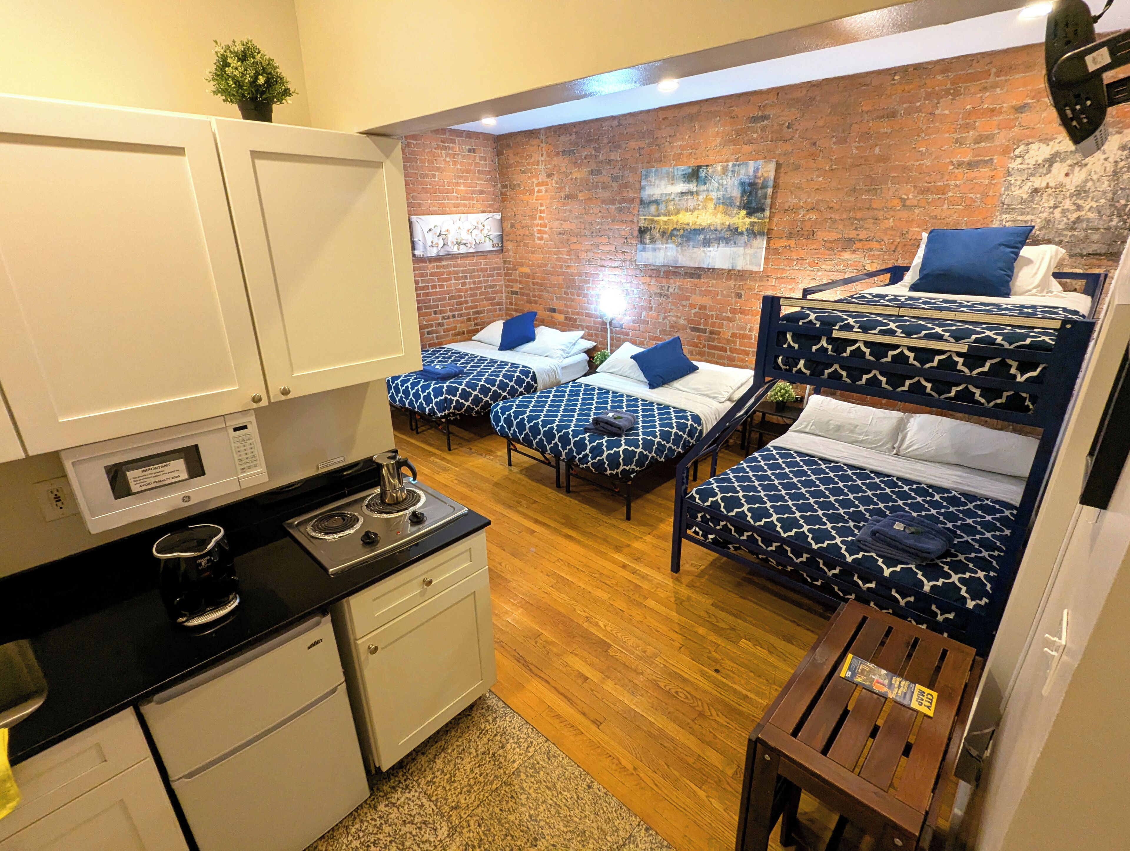 Habitación de hotel compacta con cocina, dos camas con ropa de cama estampada, una pared de ladrillo y una pequeña mesa.