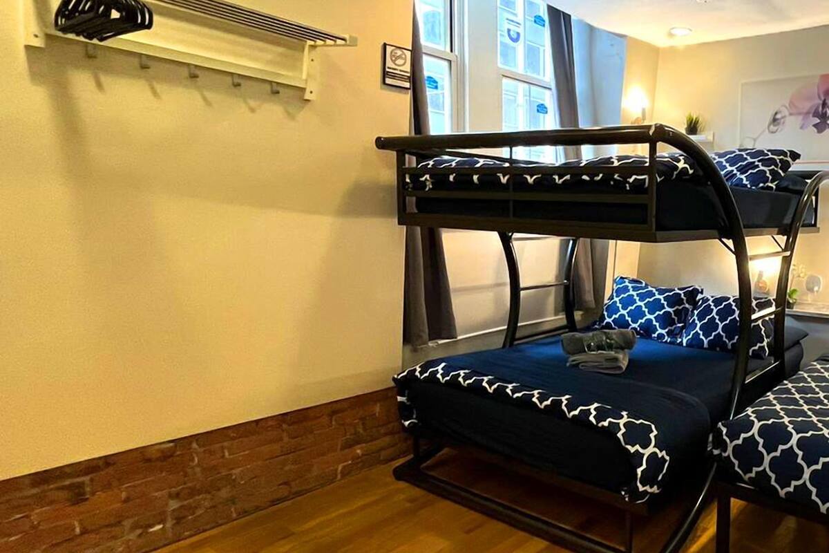 Ein Schlafraum mit Etagenbetten, einer Wand aus Backsteinen, einem Deckenventilator und einem Fenster mit Jalousien.