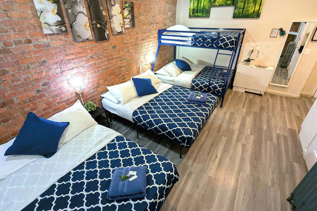 Ein Zimmer mit drei Etagenbetten, jedes mit einer gemusterten Tagesdecke und Kissen. An der rechten Seite befindet sich eine Ziegelwand und ein Spiegel.