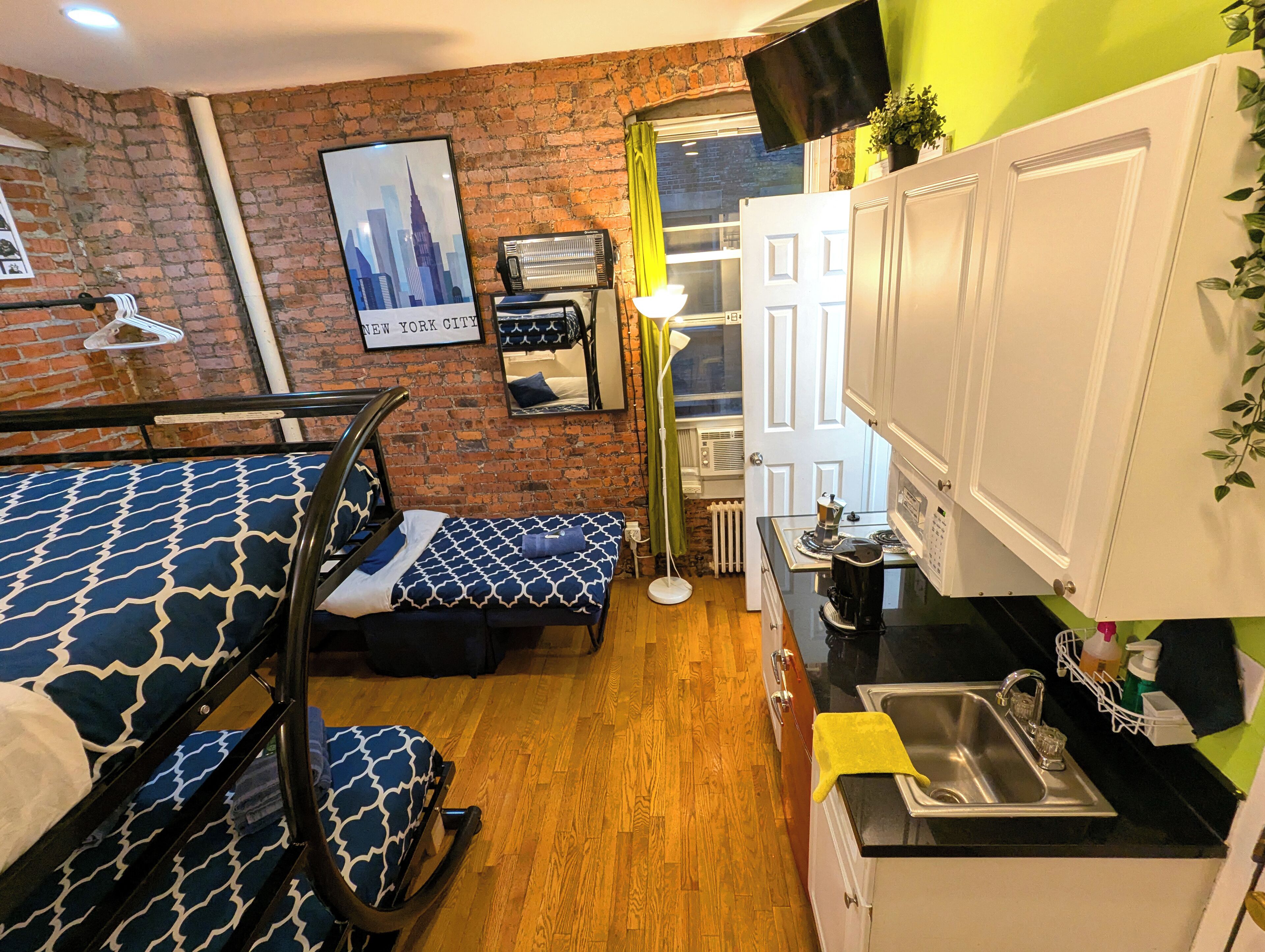 Habitación de hotel compacta con pared de ladrillo, cama con colcha estampada, pequeña kitchenette y ventana con vista al horizonte de la ciudad.