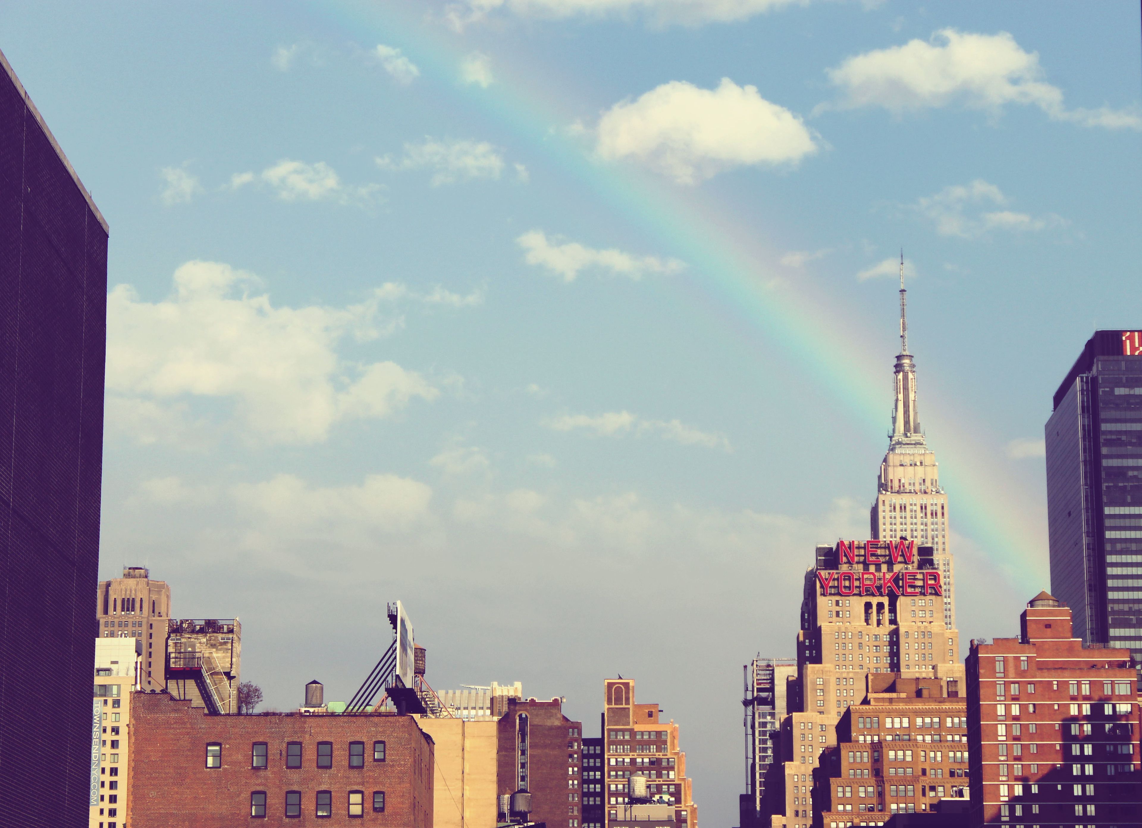 Un paisaje urbano con un edificio destacado que muestra el letrero 'NEW YORK', rodeado de otros edificios y un arcoíris visible.