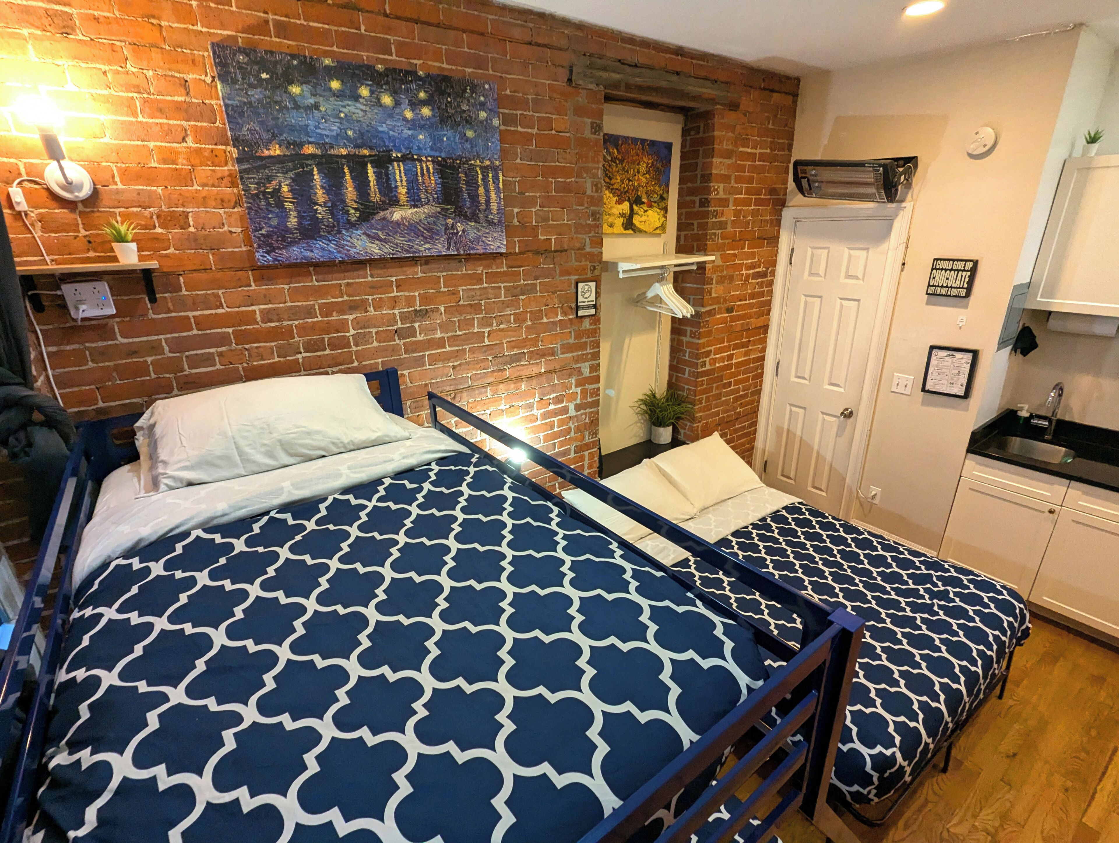 Una habitación con dos camas, pared de ladrillo, un cuadro, un espejo, una puerta y un pequeño rincón americano.