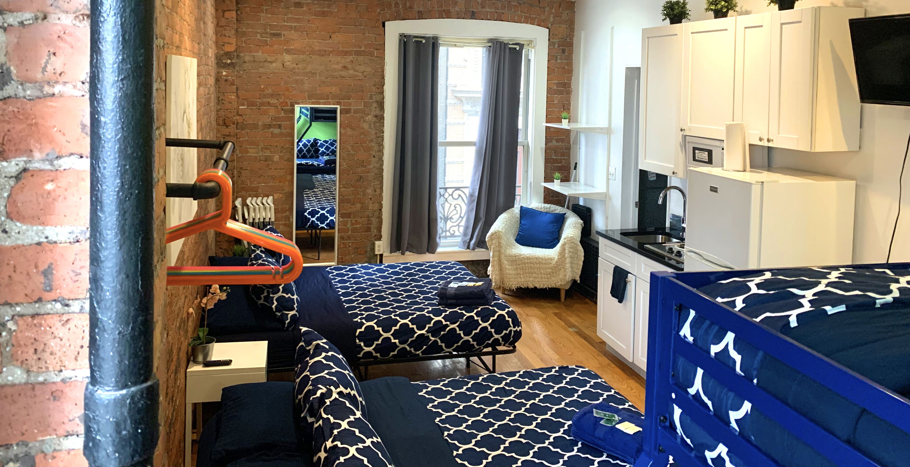 A compact room with a brick wall, a bed with patterned bedding, a chair with a cushion, a small table with a remote control, and a kitchenette area.