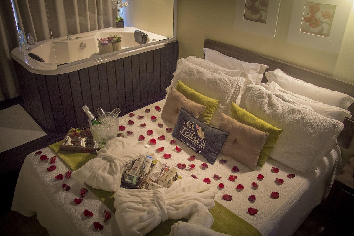 Quarto de hotel com uma banheira grande, cama bem arrumada com almofadas e um livro, e pétalas de rosa espalhadas sobre a cama.