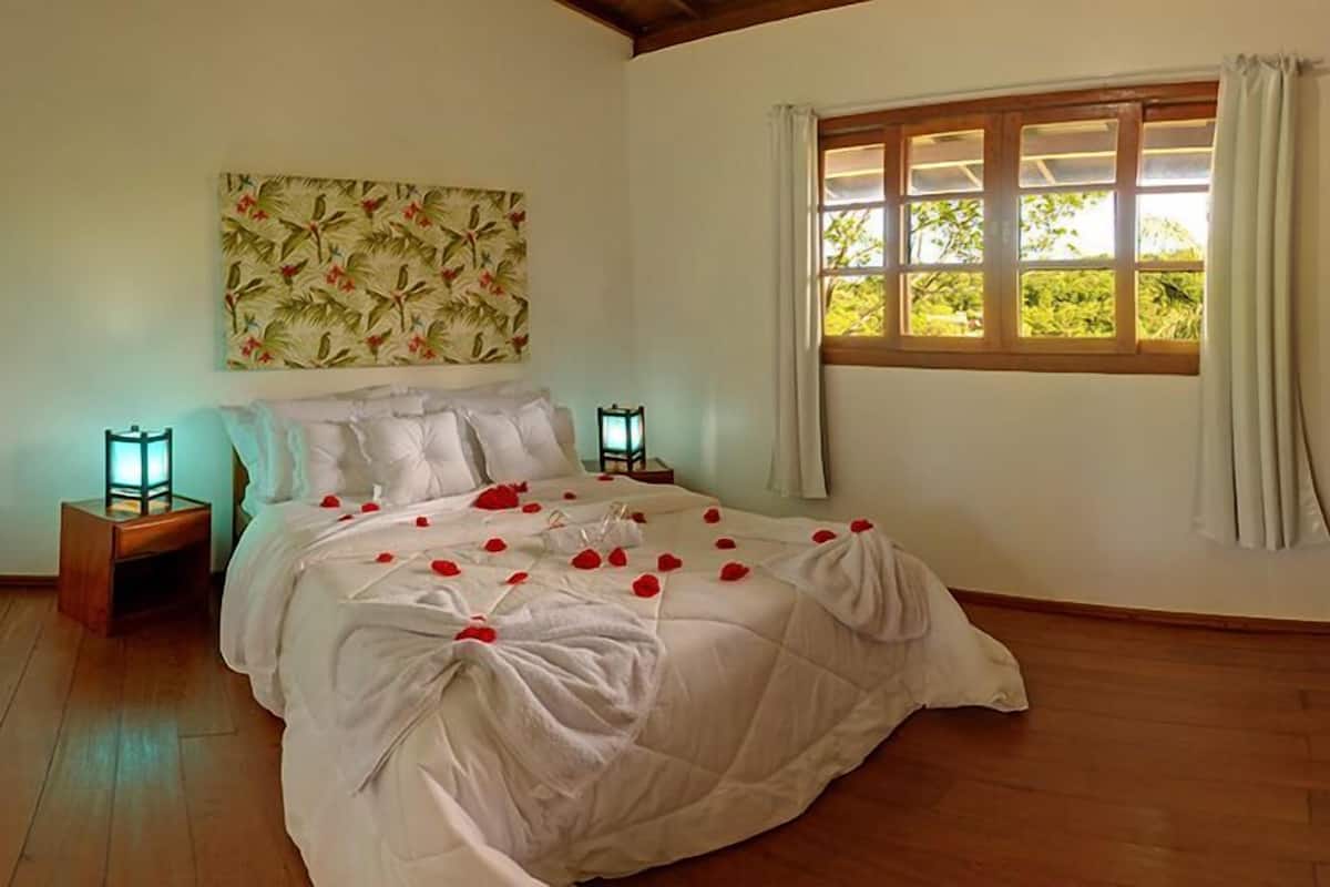 Quarto com cama, duas mesinhas de cabeceira, um criado-mudo, janela com cortinas e um painel de parede floral.
