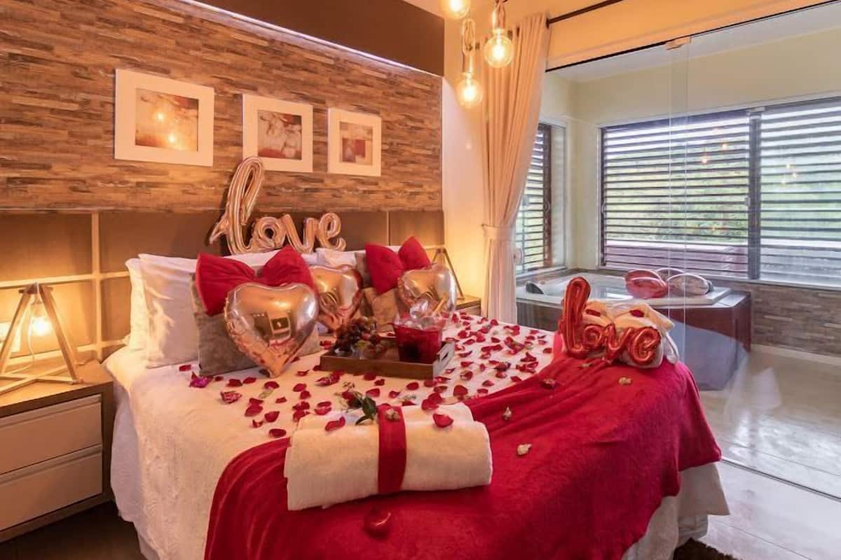 Quarto com parede de madeira, cama com roupa de cama vermelha, decorações em formato de coração e janela com persianas.