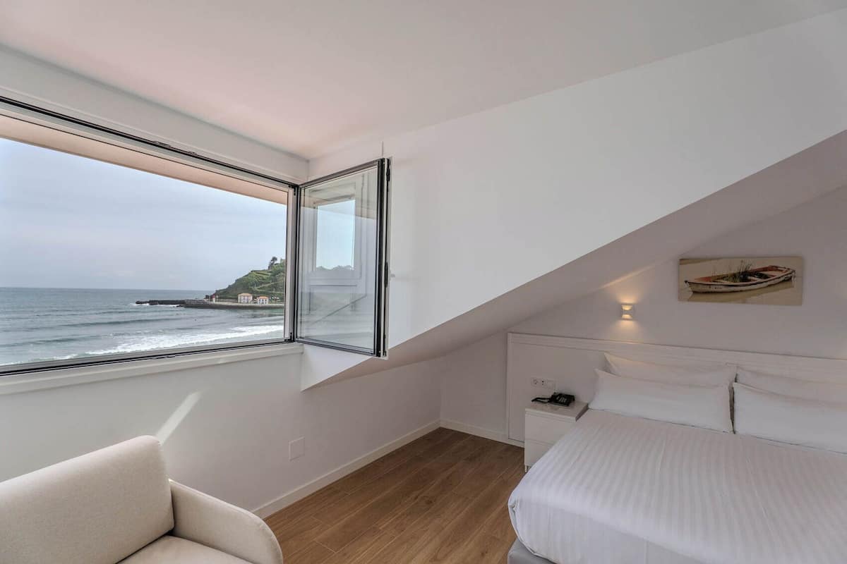 Ein Schlafzimmer mit großem Fenster und Blick auf den Ozean, einem Bett, einem Sessel und einem Nachttisch.