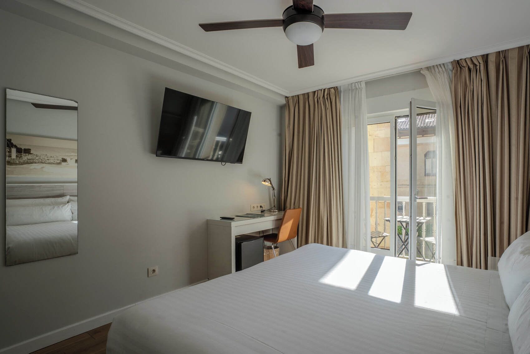 A hotel room with a bed, a desk, a chair, a TV, and a balcony with curtains.