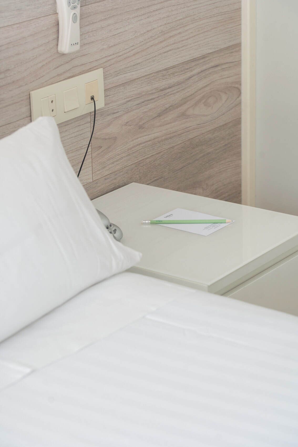 A neatly made bed with a white pillow and a green pen on a white bedside table.