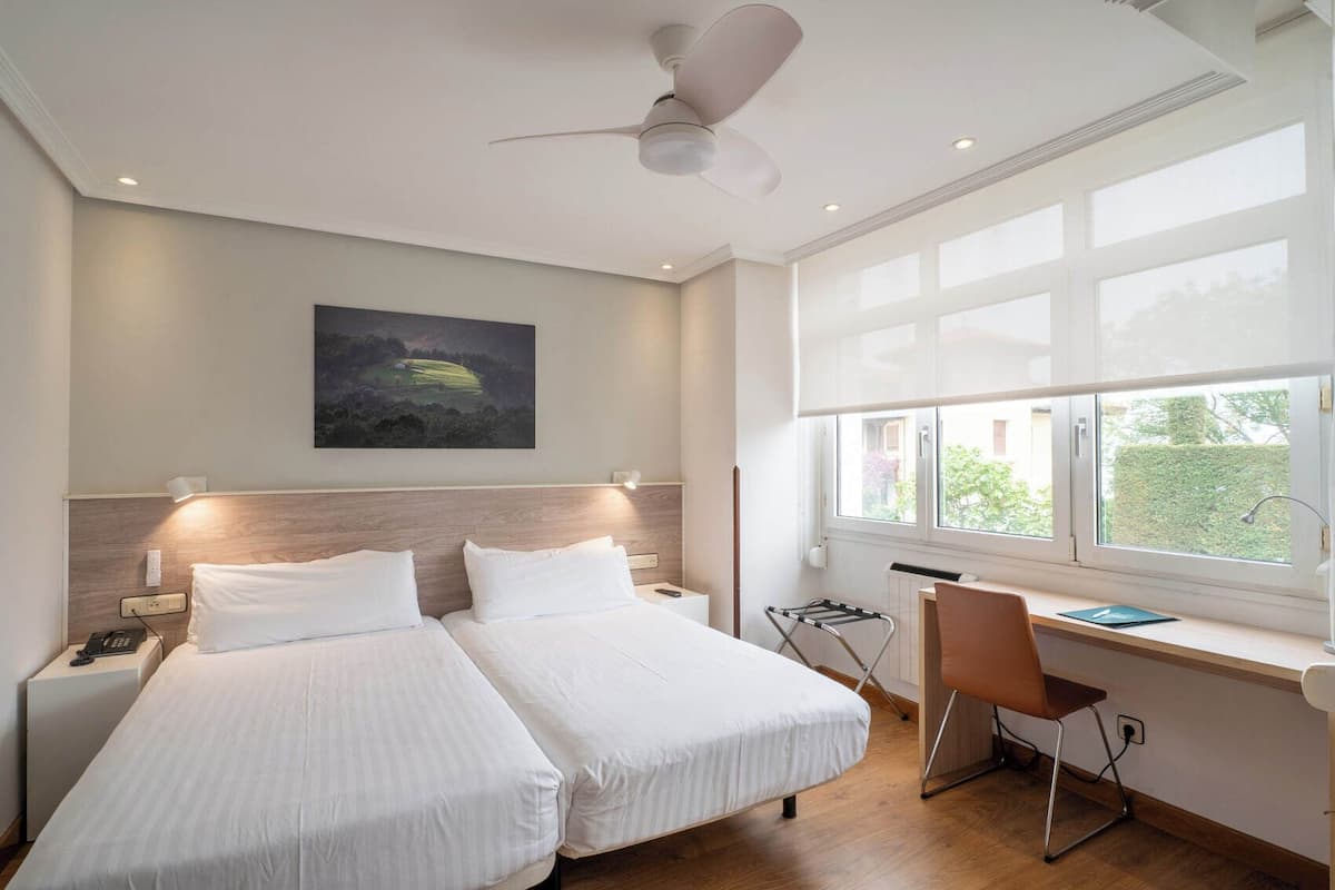 Ein Hotelzimmer mit zwei Betten, einem Schreibtisch, einem Stuhl, einem Deckenventilator, einem Gemälde an der Wand und einem Fenster mit Ausblick.