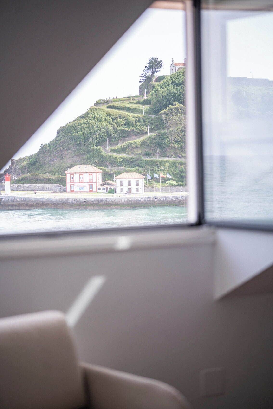 A view from a window with a building on a hill, a body of water, and a chair in the foreground.