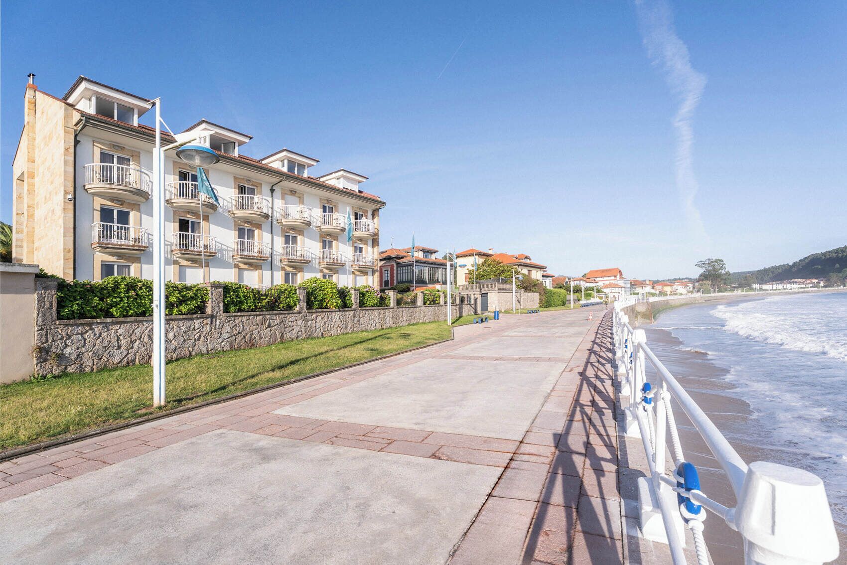 Un paseo marítimo con una hilera de edificios de varios pisos, un camino pavimentado y una barandilla con vistas al mar.