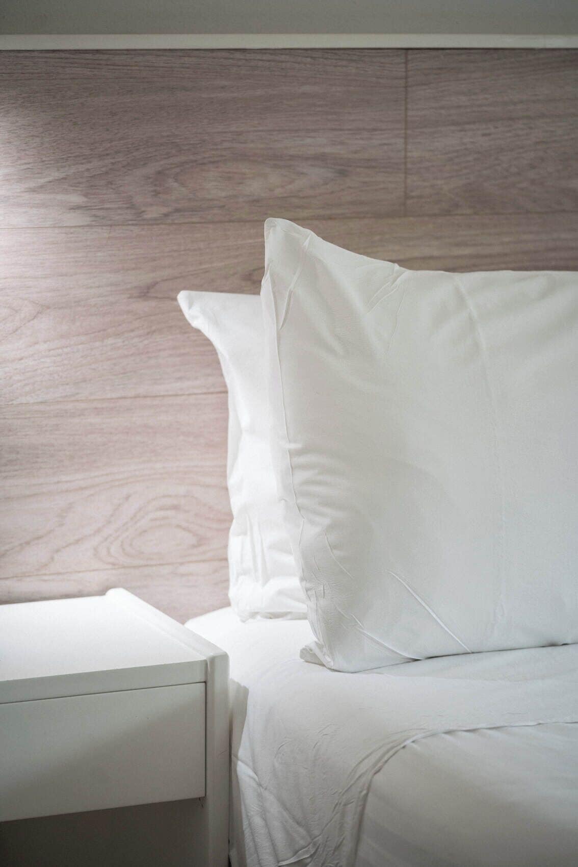 A bed with white bedding and pillows, a wooden headboard, and a white bedside table.