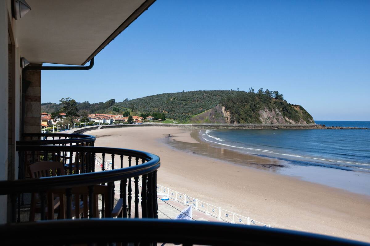 Blick auf einen Strand vom Balkon mit Geländer, ein klarer Himmel und eine ferne Hügelkette.