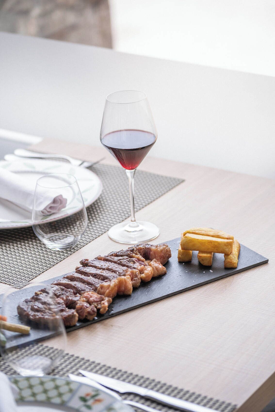 Una mesa de comedor con un bistec, una copa de vino tinto y un gofre pequeño.