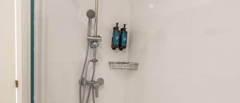 Cabina de ducha con paredes de vidrio, un cabezal de ducha manual, un grifo empotrado y un pequeño estante con artículos de tocador.
