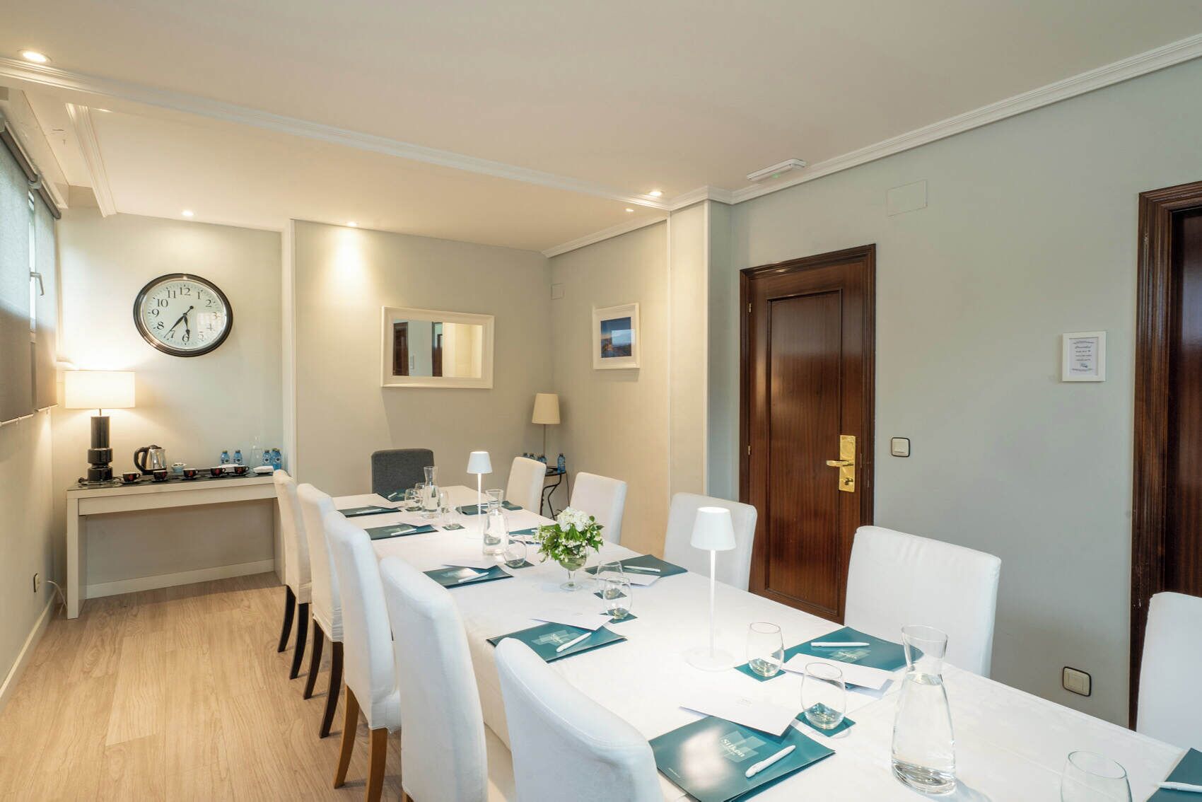 Un comedor con una mesa larga dispuesta para una comida, un reloj en la pared y una puerta con pomo.